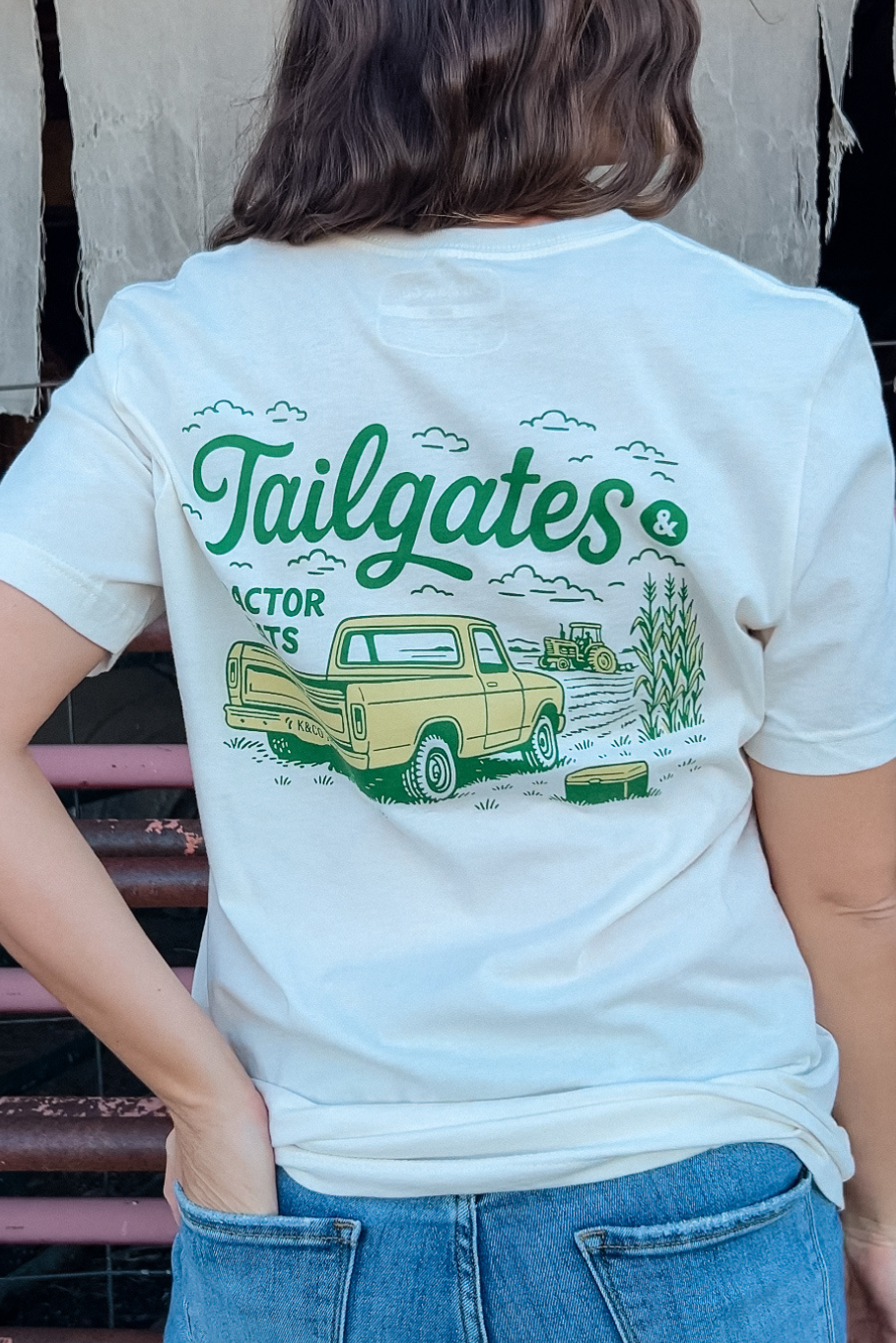 girl standing in front of cattle panels with her hand in her pocket, showing the back of her t-shirt that has a vintage pick up truck and a tractor working the field and says &quot;tailgates &amp; Tractor seats&quot;