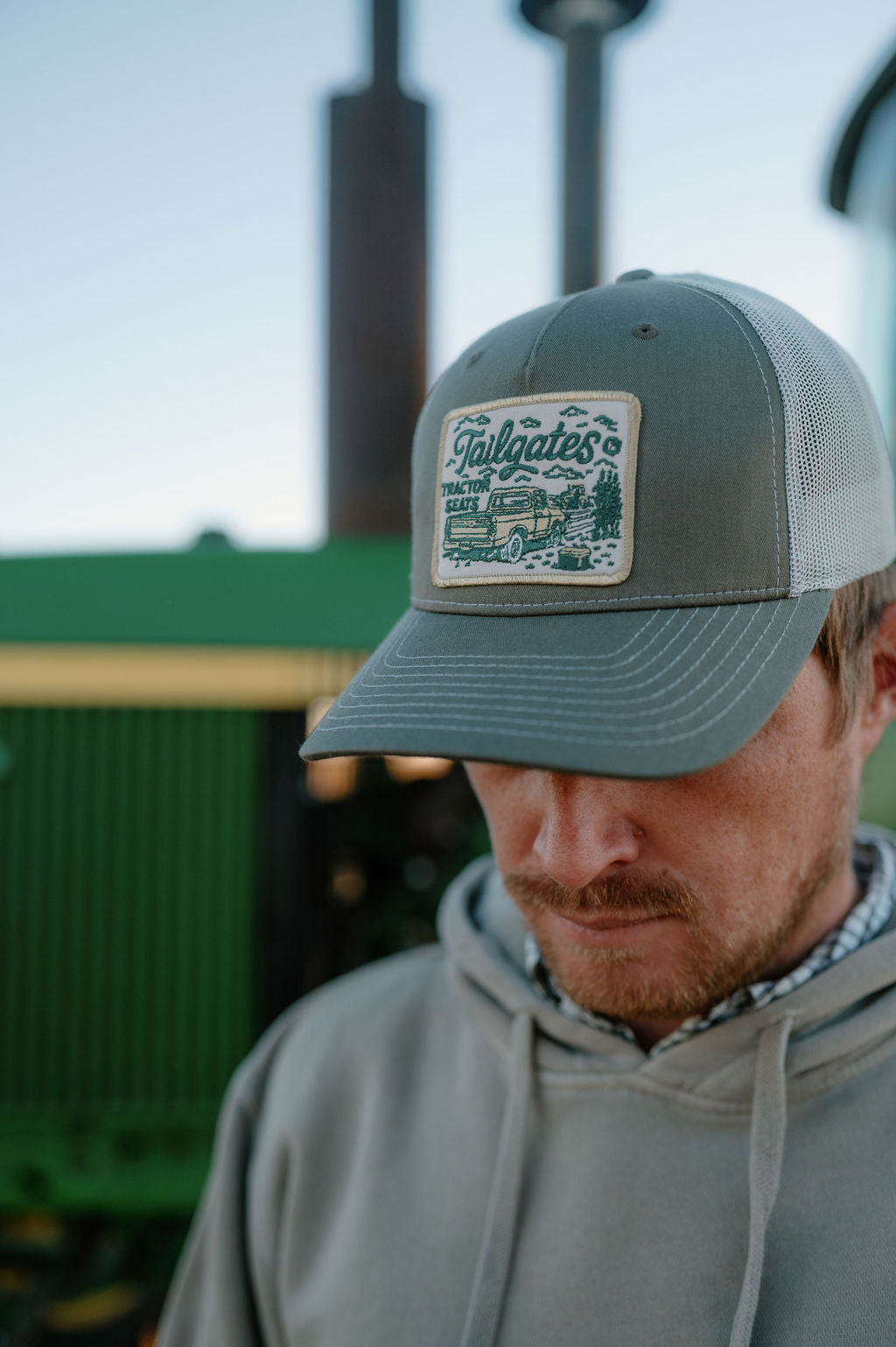 guy in front of a tractor wearing a gray blue cap with a patch that says tailgates and tractor seats
