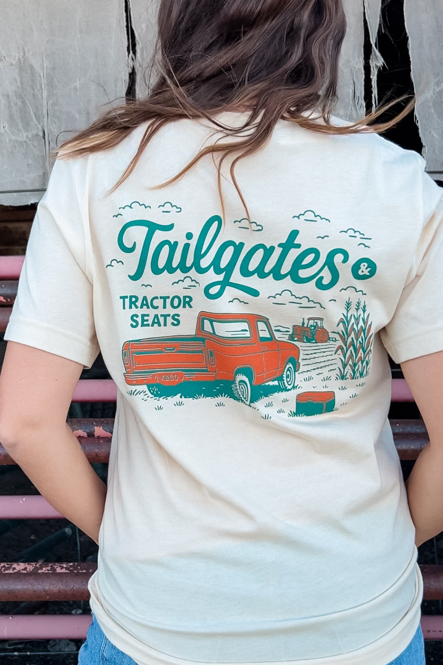 girl standing in front of cattle panels with her hand in her pocket, showing the back of her t-shirt that has a vintage pick up truck and a tractor working the field and says &quot;tailgates &amp; Tractor seats&quot; in teal and orange