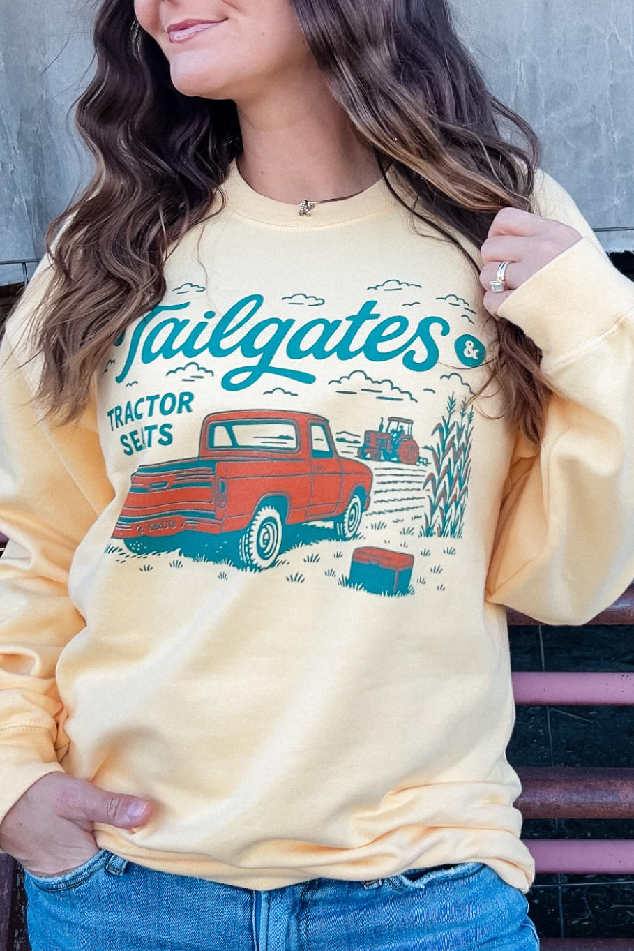 girl wearing a light yellow crewneck on a farm that has a vintage pick up truck and a tractor working a field. It says tailgates and tractor seats
