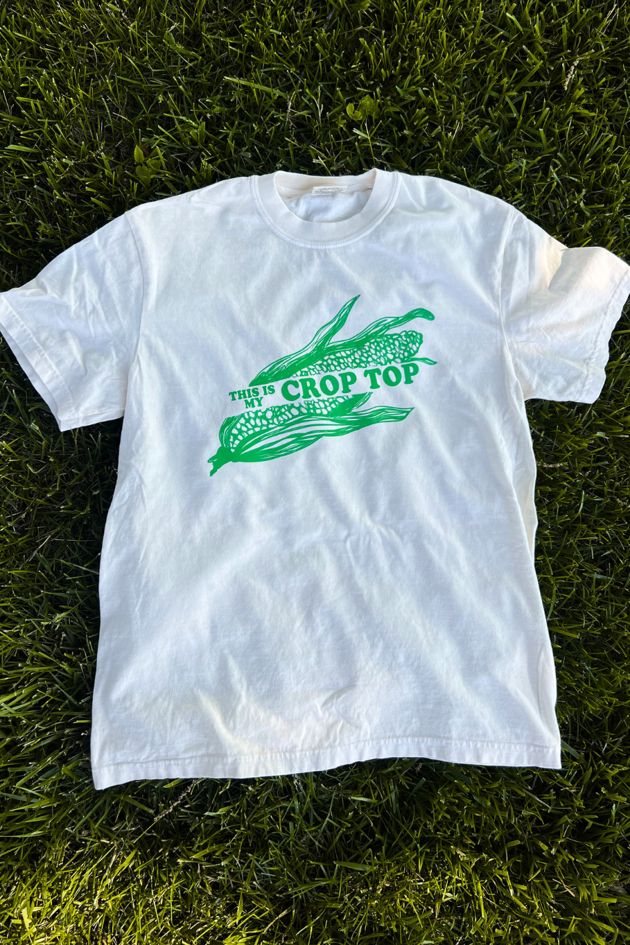flat lay of comfort colors ivory tshirt with a green graphic featuring an ear of corn with the words "this is my crop top"