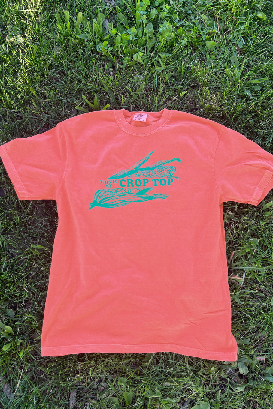 flat lay of a comfort colors tee in bright salmon with a green graphic featuring an ear of corn with the phrase " this is my crop top"