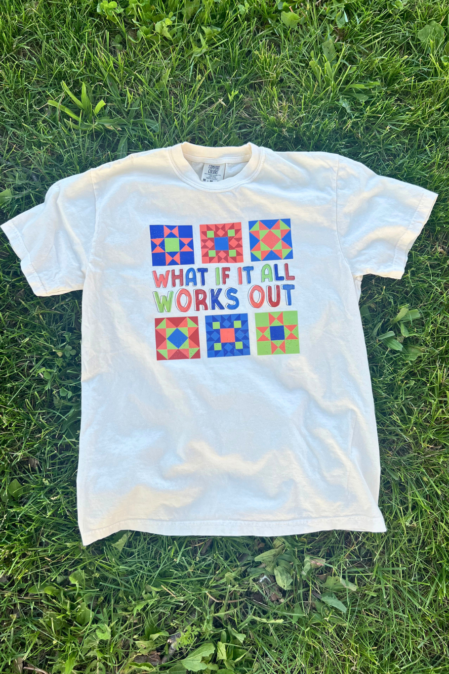 ivory graphic tshirt with colorful barn quilts and the text "what if it all works out" laying on grass
