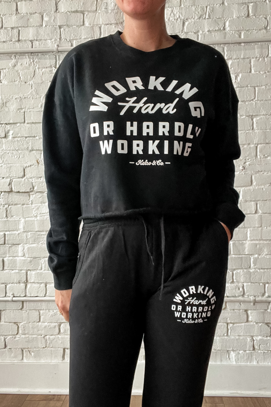 woman wearing a black cropped crewneck and joggers set that says working hard or hardly working in white