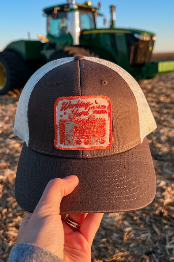 richardson 112 chocolate chip hat with orange and beige embroidered patch that says "working hard or hardly working" in front of a john deer tractor in a corn field post harvest