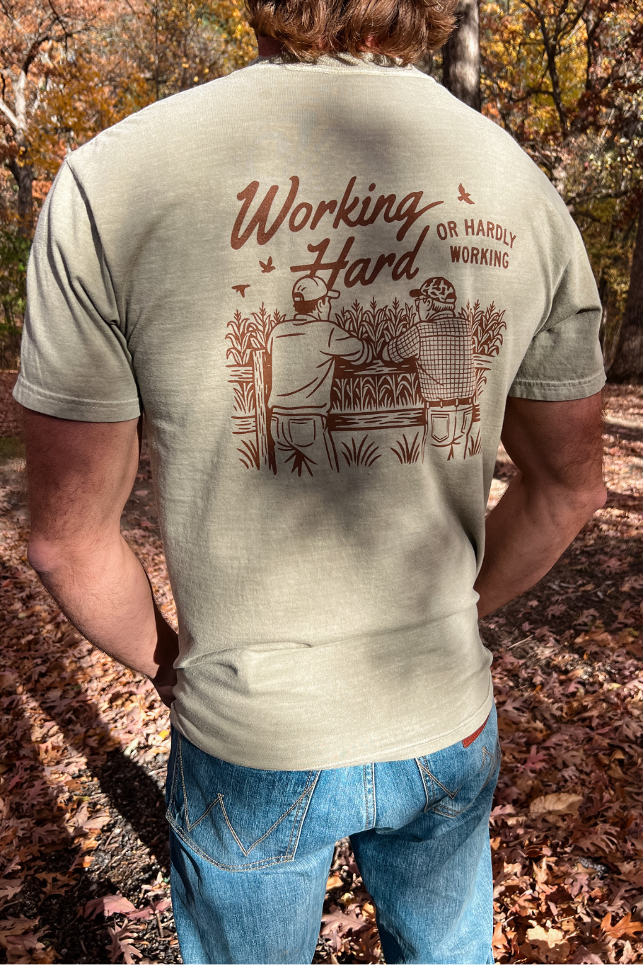 back of guy wearing comfort colors khaki shirt that says working hard or hardly working with a graphic of two farmers leaning up against a fence overlooking a cornfield