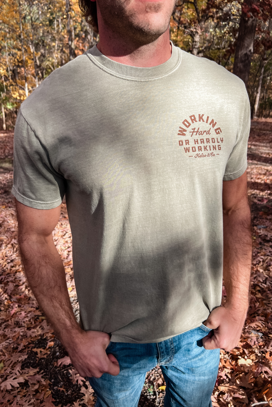 front of a guy wearing a comfort colors khaki tshirt with a graphic on the left chest that says working hard or hardly working in brown with the kelso and co logo